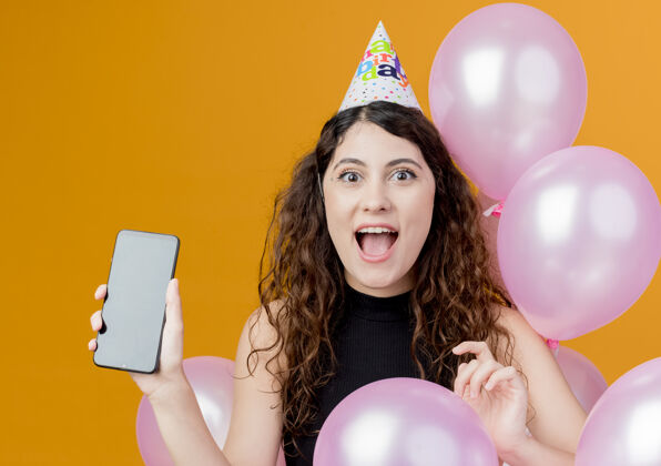 卷发一位年轻漂亮的女士 一头卷发 戴着节日帽 手持气球站在橙色的墙上 展示着智能手机的快乐和激动的生日派对理念气球年轻兴奋