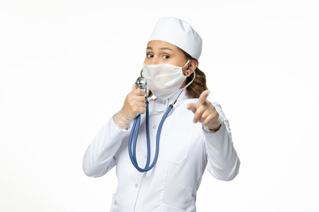 疾病正面图年轻女医生戴防护无菌口罩因冠状病毒使用听诊器在白色办公桌上大流行专业人员使用