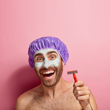 垂直高兴的年轻人用剃须刀剃胡子 涂粘土面膜 戴浴帽 有卫生和美容的程序 关心自己 光着身子 露出洁白的牙齿人类牙关面部