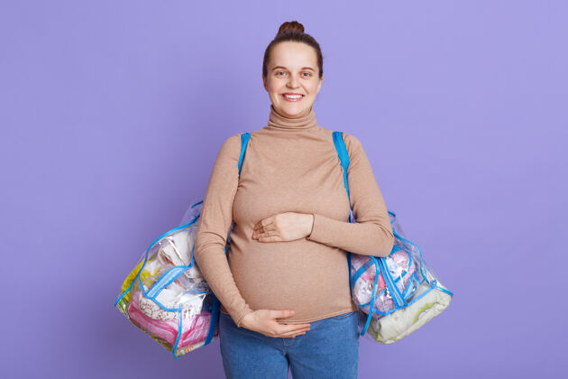 女性年轻漂亮的孕妇孤零零地站在淡紫色的墙上 摸着她的肚子 手里拿着装产房用品的袋子期待东西年轻