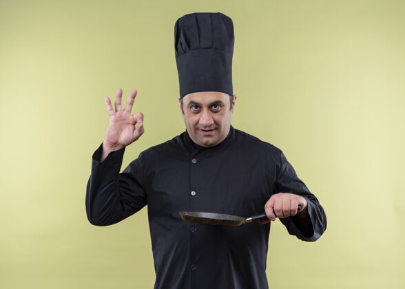 厨师男厨师身穿黑色制服 头戴厨师帽 手拿煎锅 脸上挂着ok标志 微笑着站在绿色背景上男性标志手持