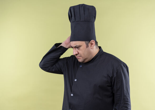 烹饪男厨师长身穿黑色制服 头戴厨师帽 困惑地看着一旁 忘记了站在绿色背景上的东西看忘了厨师