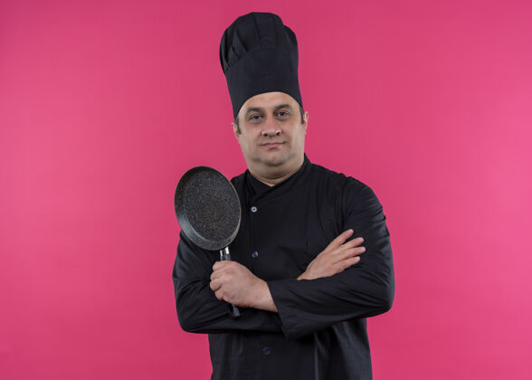 帽子男厨师身穿黑色制服 头戴厨师帽 手持平底锅 站在粉色背景下 自信地看着镜头穿着站着表情
