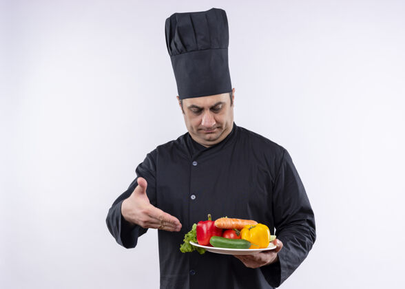 穿男厨师身穿黑色制服 头戴厨师帽 手持盛有新鲜蔬菜的盘子 手放在白色背景上帽子手臂立场