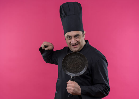 男男厨师身穿黑色制服 头戴厨师帽 手持平底锅 持刀威胁 面带微笑地看着摄像机站在粉色背景下站着烹饪黑色