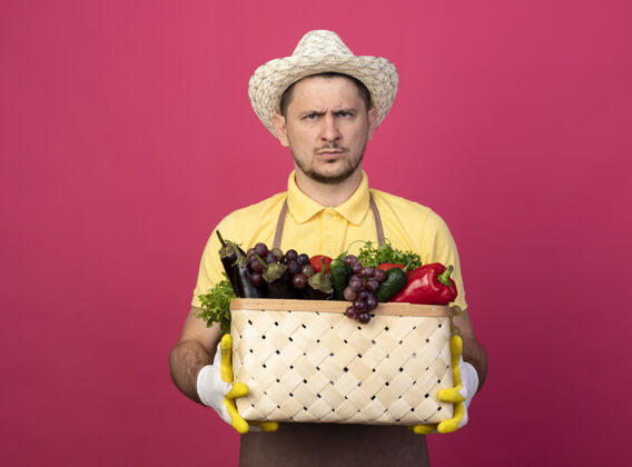 手套年轻的园丁戴着工作手套 穿着连体衣 戴着帽子 手里拿着装满蔬菜的箱子 愁眉不展不高兴蔬菜皱眉