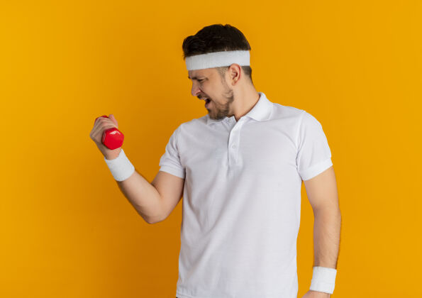 紧张身穿白衬衫 头戴钢带的年轻健身男子站在橙色背景上练习哑铃 看起来很紧张看站健身
