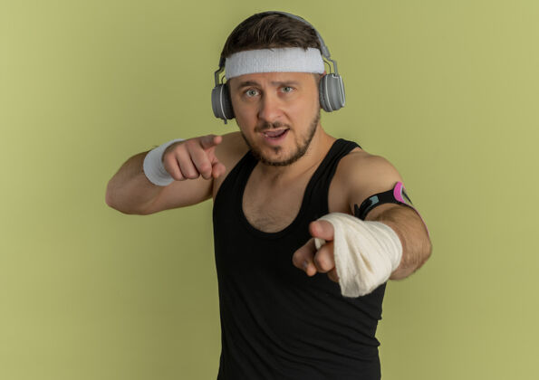 耳机戴着头带和耳机的年轻健身男子站在橄榄色背景下 戴着智能手机袖标看着摄像机训练男人健身臂带