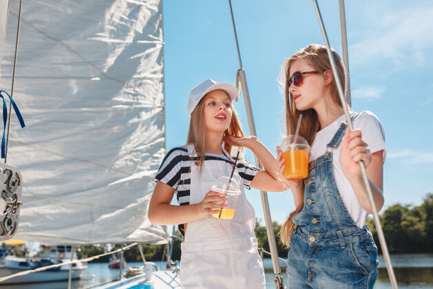 欢呼孩子们在游艇上喝着橙汁青少年或少女们在蓝天下户外五颜六色的衣服孩子们的时尚 阳光明媚的夏天 河流和假日的概念年轻人姐妹自然