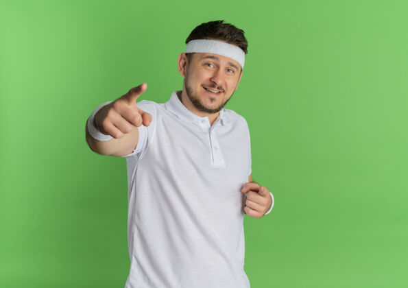 手指身穿白衬衫 头箍 手指对着相机 脸上带着微笑的年轻健身男子站在绿色背景下男人脸手指