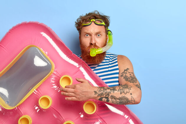 条纹照片中的红发姜男戴着泳镜 戴着潜水面罩 要在海里潜水游泳 手里拿着粉色充气床垫 神情严肃水平假期卷发