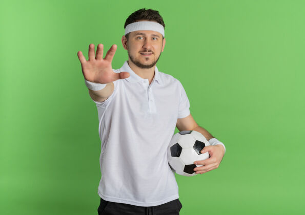 白色身穿白衬衫 头戴足球带的年轻健身男子举着双臂站在绿色背景上看着摄像机头带足球手臂