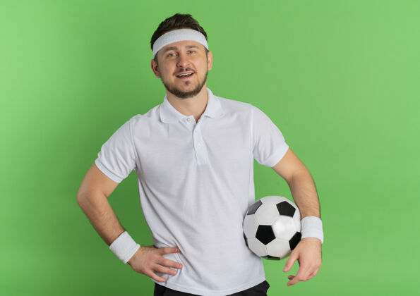 白色身穿白衬衫 头戴足球带的年轻健身男子站在绿色背景下 兴高采烈地微笑着看着摄像机绿色欢呼足球