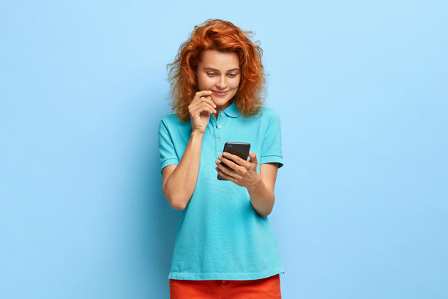 成人心满意足的可爱姜女手持手机 喜闻乐见 穿着休闲服 站在蓝色的墙壁上 享受使用现代科技 微笑温柔电话短信年轻人
