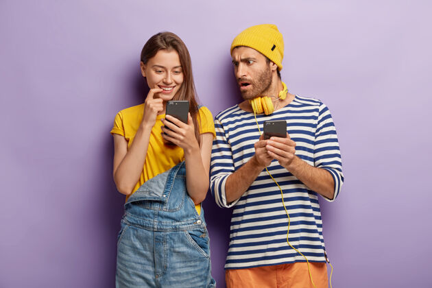 工作服千禧年的女孩积极地看着智能手机 用手机惊呆了迷惑不解的家伙 紧紧地站在紫色的墙壁上年轻的现代科技沉迷的情侣情侣年轻通信