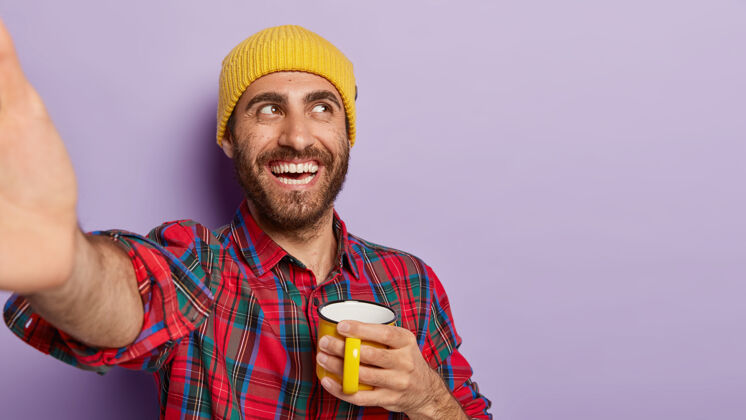 杯子开朗的没刮胡子的男生喜欢喝黄色杯子里的热饮 自拍 伸开胳膊 积极地微笑 戴着黄色帽子和格子衬衫时尚紫色人