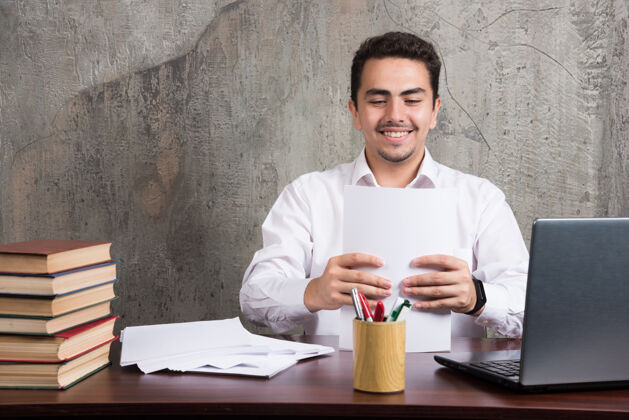 笔记本电脑微笑的男人拿着几张纸坐在桌子旁高质量的照片男人员工桌子