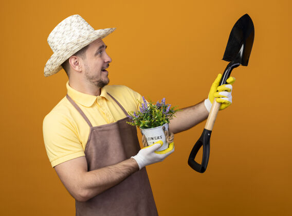 人穿着连体衣 戴着帽子的年轻园丁拿着铲子和盆栽植物看着铲子 站在橙色的墙上 脸上带着幸福的笑容抱着站着植物