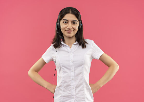 年轻人身穿白衬衫 戴着耳机的年轻女孩站在粉色的墙上 面带微笑 自信地站在前面耳机女孩表情