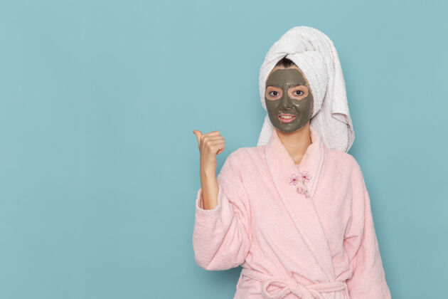 面霜正面图身着粉色浴袍的年轻女性 脸上戴着面具 蓝色墙壁上的淋浴清洁美容自护霜服装面具前面
