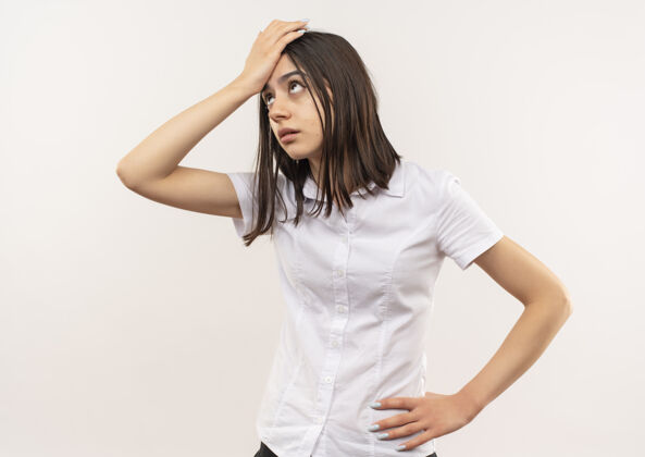衬衫穿白衬衫的年轻女孩一边看一边困惑地用手捂着头 因为她站在白墙上犯了错误姿势女人人