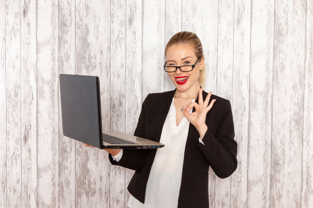 女商人正面图穿着严苛衣服的年轻女商人黑色夹克用她的笔记本电脑在白墙上眨眼上班上班上班的女商人眨眼前台使用