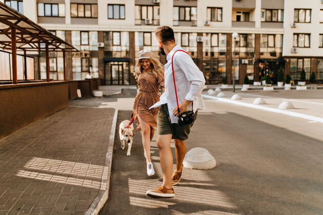 家庭穿着夏季服装的时尚夫妻在公寓的背景上和他们的狗一起奔跑和玩耍穿着浅色衬衫的男人牵着他心爱的手 拿着相机红领人圆点连衣裙