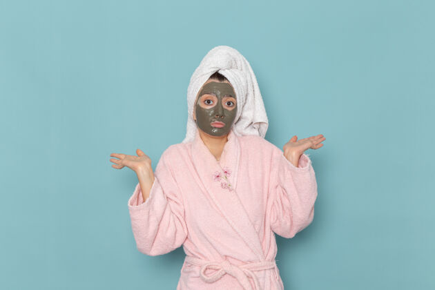 脸正面图身着粉色浴袍 脸上戴着面具的年轻女性在蓝色墙壁上摆姿势沐浴清洁美容自护霜伪装人姿势