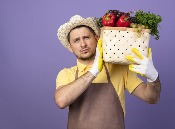 人年轻的园丁穿着连体衣 戴着帽子 戴着工作手套 肩上扛着装满蔬菜的箱子 站在紫色的墙上显得很自信满满的箱子拿着