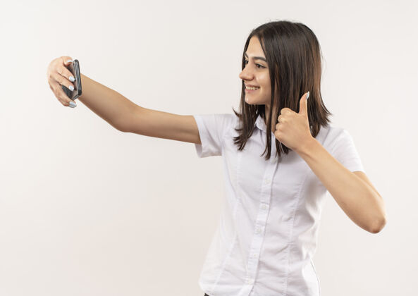 公民穿着白衬衫的年轻女孩用手机自拍 微笑自信地站在白墙前竖起大拇指拇指站立人
