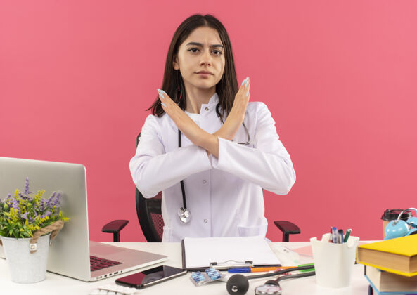 坐着年轻的女医生穿着白大褂 脖子上戴着听诊器 严肃地做着停车标志 双手交叉着脸坐在桌旁 笔记本电脑放在粉红色的墙上笔记本电脑桌子女性