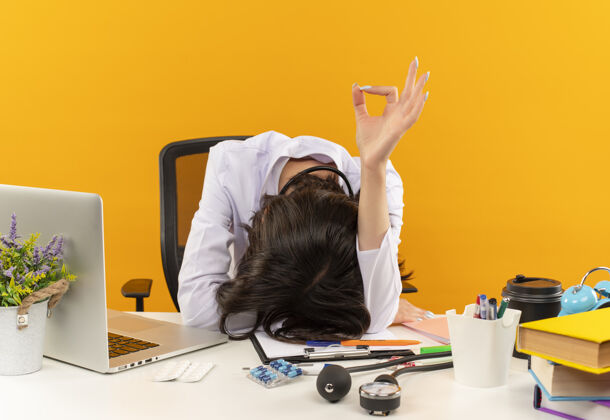 表演身穿白大褂的年轻女医生躺在桌子上 手里拿着笔记本电脑和文件 橙色的墙上写着“ok”的牌子医疗保健办公室工作