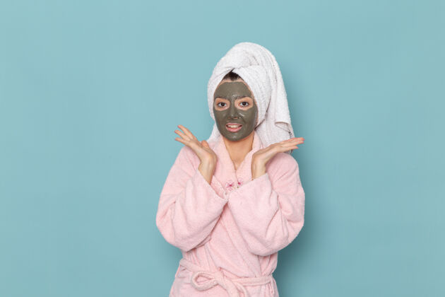 伪装正面图：年轻女性 穿着粉色浴袍 沐浴后带着面具微笑 在蓝色墙壁上 美丽的水霜 自我护理淋浴浴室淋浴前面肖像