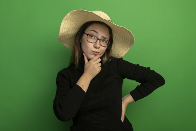 站着年轻漂亮的女人戴着夏天的帽子 穿着黑色高领毛衣 戴着眼镜 带着怀疑的表情 手放在下巴上 站在绿色的墙上思考怀疑下巴年轻