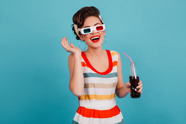 情绪幽默的小女孩戴着3d眼镜摆姿势摄影棚拍摄了一个幸福的黑发女人 蓝色背景上隔着一瓶苏打水发型女性肖像