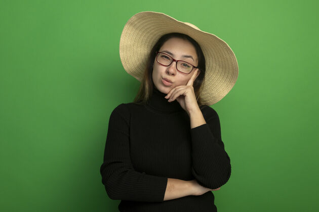姿势年轻漂亮的女人戴着夏天的帽子 穿着黑色高领毛衣 戴着眼镜 带着怀疑的表情站在绿色的墙上看着前面怀疑穿着女人