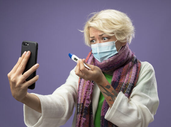 围巾有病的不健康的妇女 短头发 戴着暖和的围巾 戴着面罩 拿着温度计 在紫色的墙上用手机通话女人面具生病
