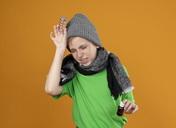 痛苦生病的小男孩穿着绿色t恤 戴着暖和的围巾和帽子 感觉不舒服 手里拿着药瓶和药丸 头痛难受 站在橙色的墙上不开心站着药丸小