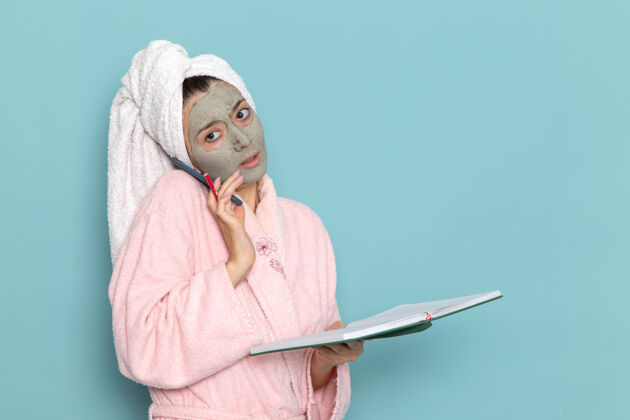 男性正面图身着粉色浴袍的年轻女性淋浴后在蓝色墙壁上打电话美容水霜自理淋浴脸前面后面