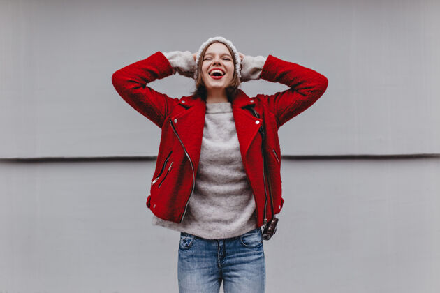 奢侈品穿着红色夹克 浅色牛仔裤和羊绒衫的女孩戴上帽子 对着白墙大笑衣服温暖套装