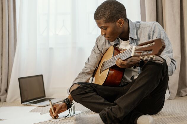 乐器手男性音乐家在床上和笔记本电脑上用吉他写音乐的前视图人才房子创意