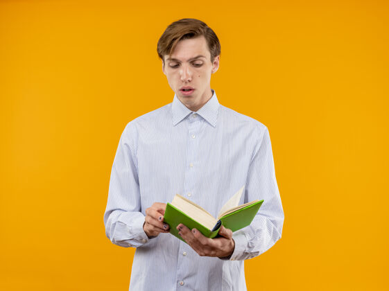 严肃身穿白衬衫的年轻人拿着打开的书 严肃地站在橙色的墙上看着书站着年轻男人