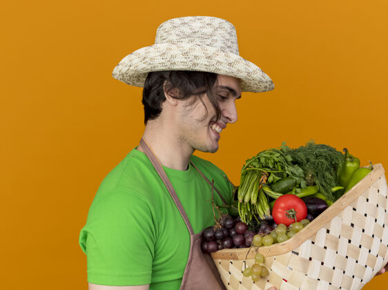 看着年轻的园丁 围着围裙 戴着帽子 手里拿着装满蔬菜的箱子 站在橙色的墙上 面带微笑地看着它微笑花园脸