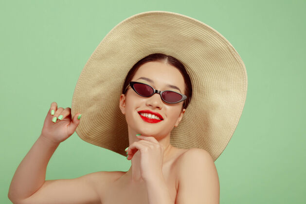 健康美丽女人的肖像 明亮的妆容 帽子和太阳镜在绿色的工作室背景上时髦和时尚的制作和发型夏天的颜色美丽 时尚和广告概念微笑年轻太阳镜严肃