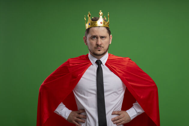 看穿红斗篷的超级英雄商人戴着王冠 严肃的脸 胳膊放在臀部 站在绿色的墙上皇冠穿严重