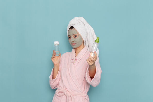奶油正面图身着粉色浴袍的年轻女性在淋浴后手持喷雾在蓝色墙壁上进行美容水霜自我护理淋浴淋浴浴袍年轻人