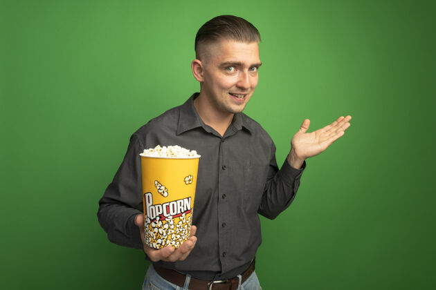 介绍身穿灰色衬衫的年轻帅哥拿着装满爆米花的水桶望着前方 微笑着举起手臂 在绿色的墙上展示着什么衬衫绿色提高