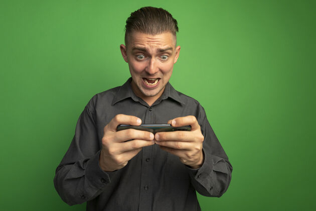 人穿着灰色衬衫的年轻帅哥用智能手机玩游戏兴奋而情绪激动地站在绿色的墙上人绿色面部表情