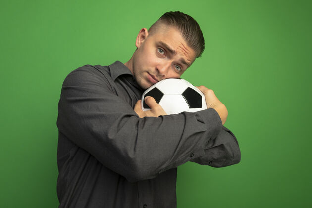 头身穿灰色衬衫的年轻帅哥抱着足球 把头靠在球上 带着悲伤的表情站在绿色的墙上看着前方足球球悲伤