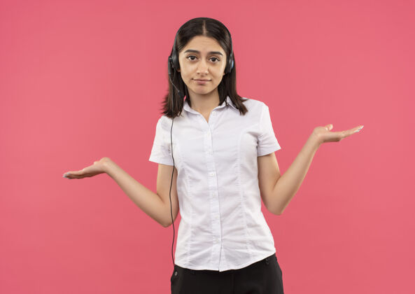 侧线穿着白衬衫戴着耳机的年轻女孩 望着前面 两臂叉开 站在粉红色的墙上衬衫耳机人